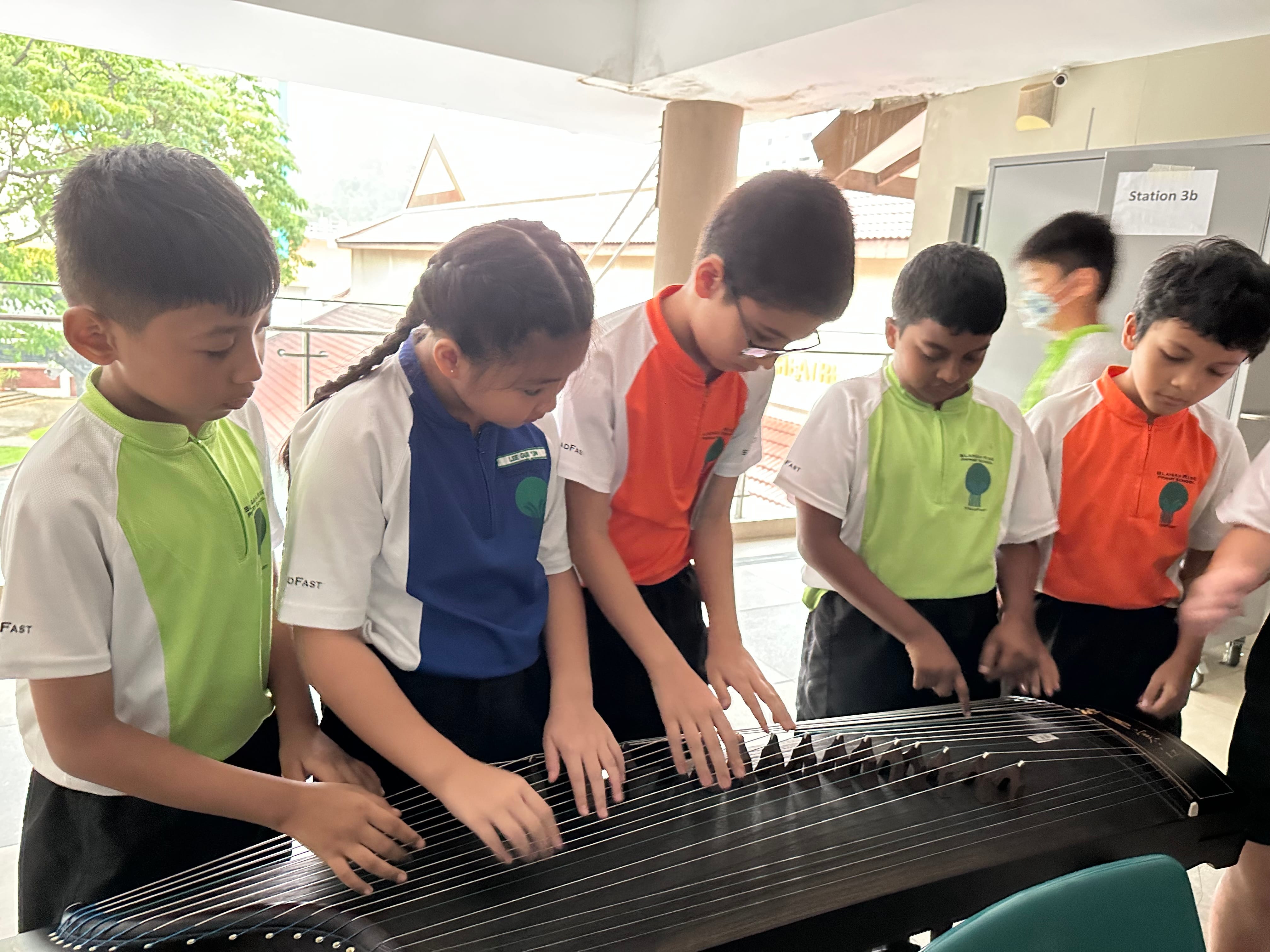 Five school children in uniforms stand around a guzheng, touching its strings outdoors at Station 3b.